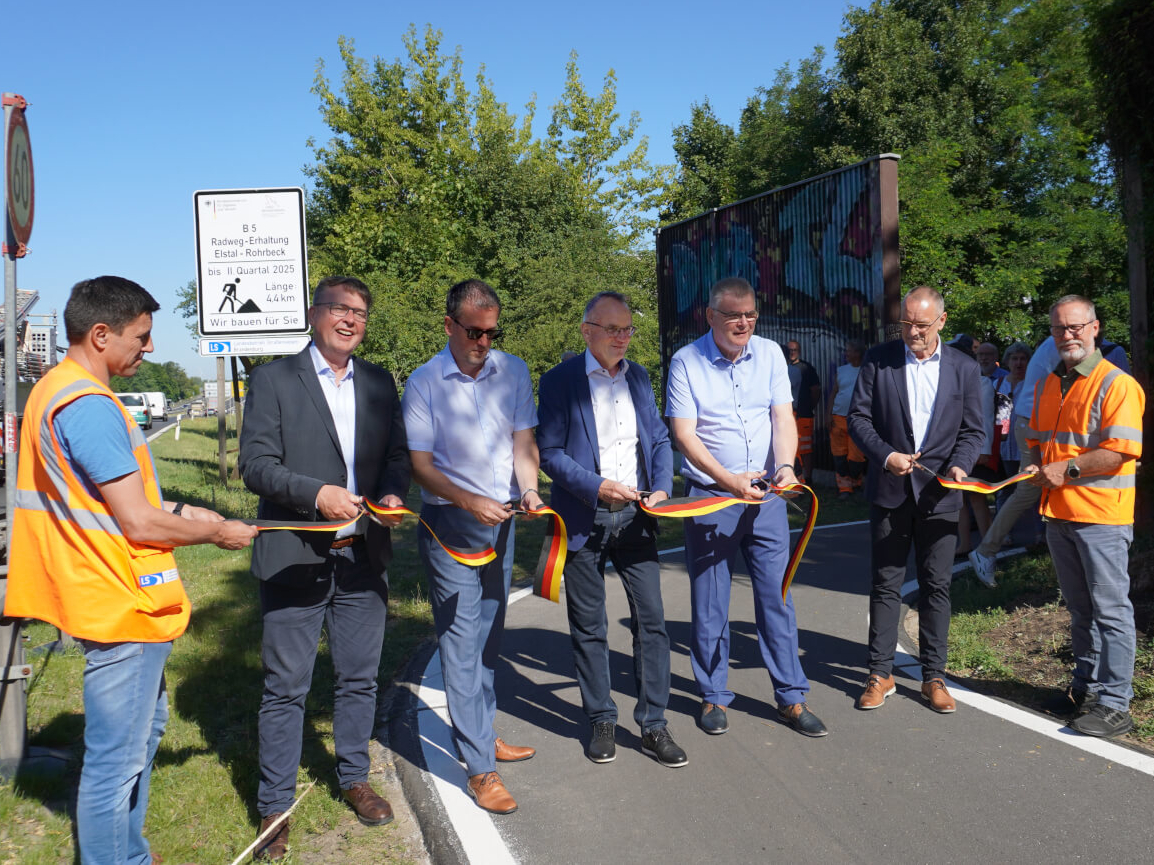 Verkehrsfreigabe RW B5 Elstal Das Band in den Farben schwarz rot gold wird zur Verkehrsfreigabe von einigen Gästen durchgeschnitten. In Hintergrund befindet sich das Baustellenschild.